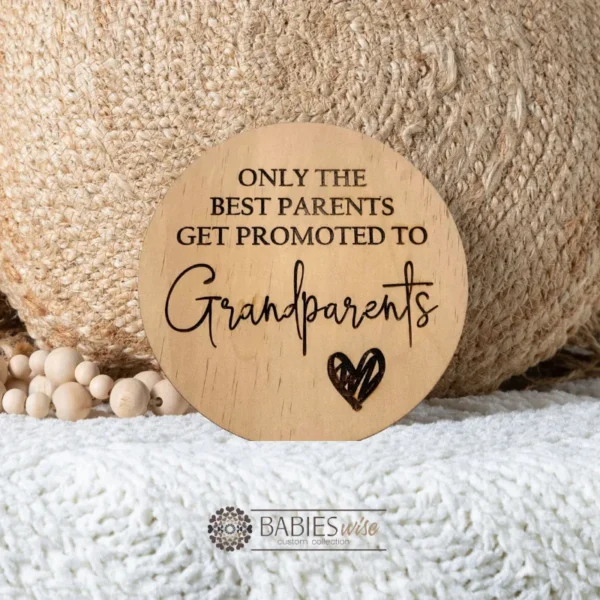 Laser-engraved round timber plaque with the message "only the best parents get promoted to Grandparents" in a mix of uppercase and script fonts, featuring a small engraved love heart at the bottom. Styled on a soft white blanket with a woven pillow and wooden beads.