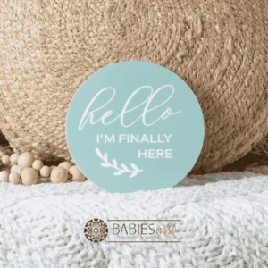 Mint round acrylic uv printed announcement plaque with white lettering reading "Hello I'm Finally Here" and a small leaf illustration.Styled on a soft white blanket with a woven pillow & wooden beads.
