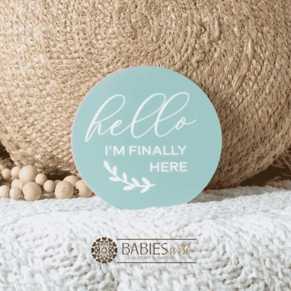 Mint round acrylic uv printed announcement plaque with white lettering reading "Hello I'm Finally Here" and a small leaf illustration.Styled on a soft white blanket with a woven pillow & wooden beads.