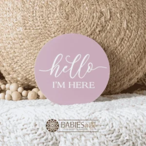 Dusty pink round birth announcement plaque with white leterung reading "Hello I'm Here.Styled on a soft white blanket with a woven pillow & wooden beads.