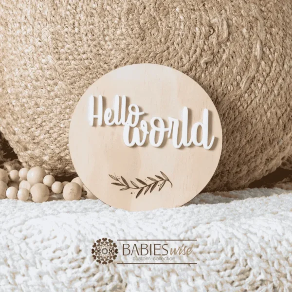 Round natural timber birth announcement plaque with raised 3D acrylic lettering reading "Hello World" and engraved leaf motif. Styled on a soft white blanket with a woven pillow & wooden beads.