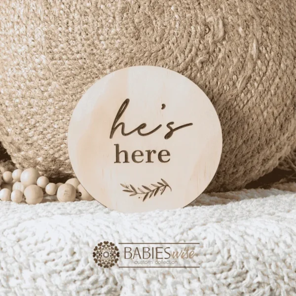 Round natural timber birth announcemnt plaque engraved with the words "he's here" and a small leaf motif.Styled on a soft white blanket with a woven pillow & wooden beads.