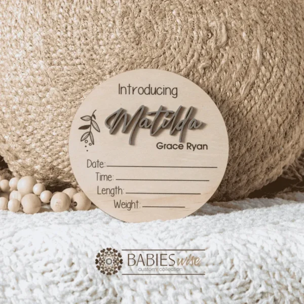 Round timber announcement plaque featuring the word "Introducing" above a personlised raised 3D acrylic name, with spaces below to record a baby's birth date, time, length, and weight. Styled on a soft white blanket with a woven pillow & wooden beads.