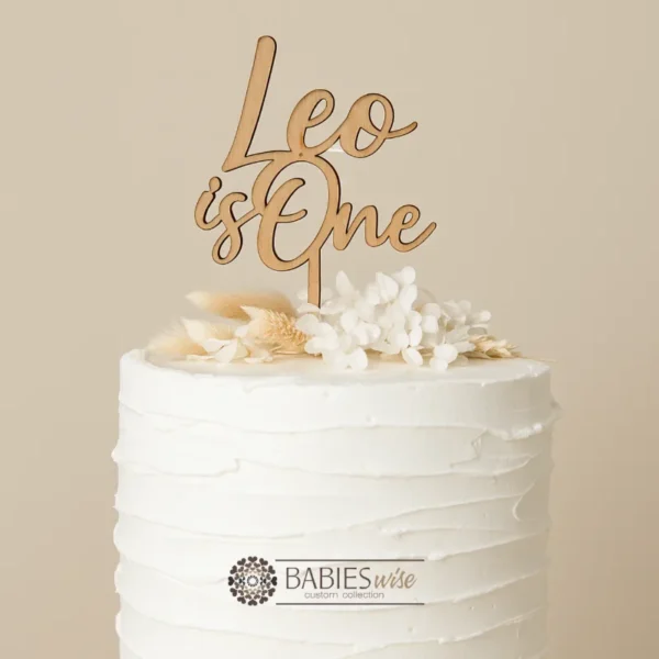 Personlised timber first birthday cake topper featuring your child's name in a script lettering above the words "is One" Styled on a white cake with a floral arrangement.