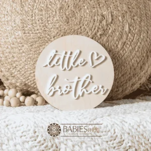 Round timber sibling announcement plaque with raised white acrylic script reading "little brother" and a small loveheart detail. Styled on a soft white blanket with a woven pillow & wooden beads.