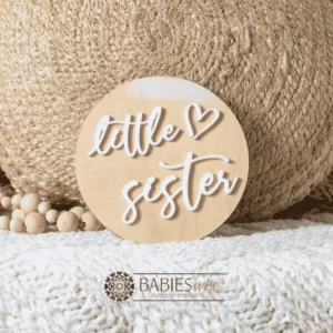 Round timber sibling announcement plaque with raised white acrylic script reading "little sister" and a small loveheart detail. Styled on a soft white blanket with a woven pillow & wooden beads.