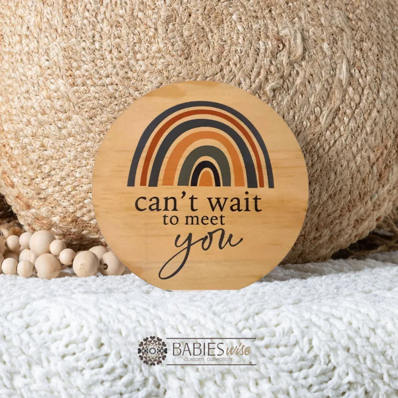Round timber pregnancy announcement plaque with blue rainbow design and "can't wait to meet you" text. Styled on soft blanket with a woven pillow and wooden beads.