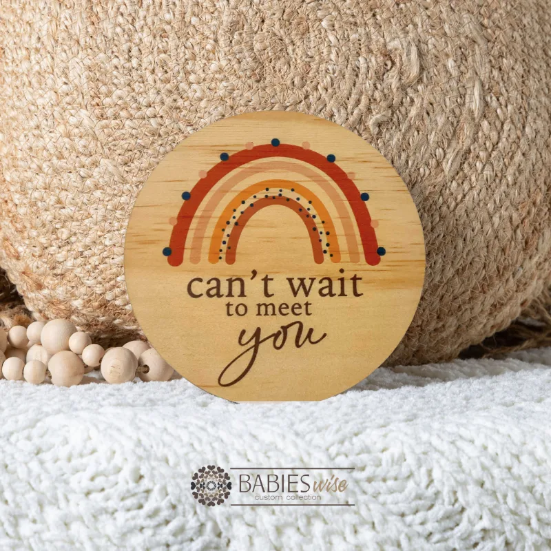 Round timber pregnancy announcement plaque with blue rainbow design and "can't wait to meet you" text. Styled on soft blanket with a woven pillow and wooden beads.