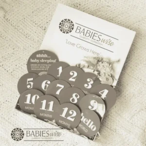 Mocha acrylic monthly milestone gift box featuring 12 round milestone discs with white 3D numbers, a matching Birth Announcement plaque, "Shh.. Baby Sleeping plaque presented in a white magnetic gift box with shredded straw. Styled on a soft cream blanket.