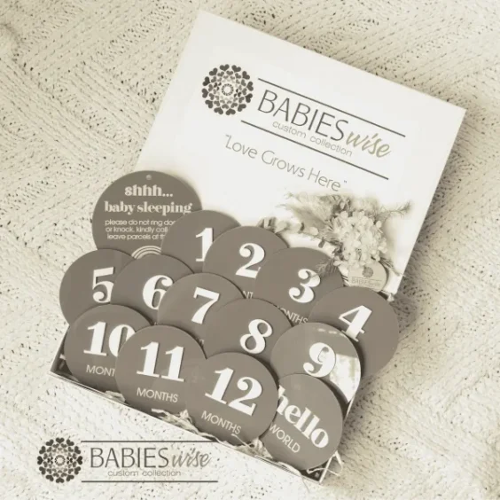 Mocha acrylic monthly milestone gift box featuring 12 round milestone discs with white 3D numbers, a matching Birth Announcement plaque, "Shh.. Baby Sleeping plaque presented in a white magnetic gift box with shredded straw. Styled on a soft cream blanket.