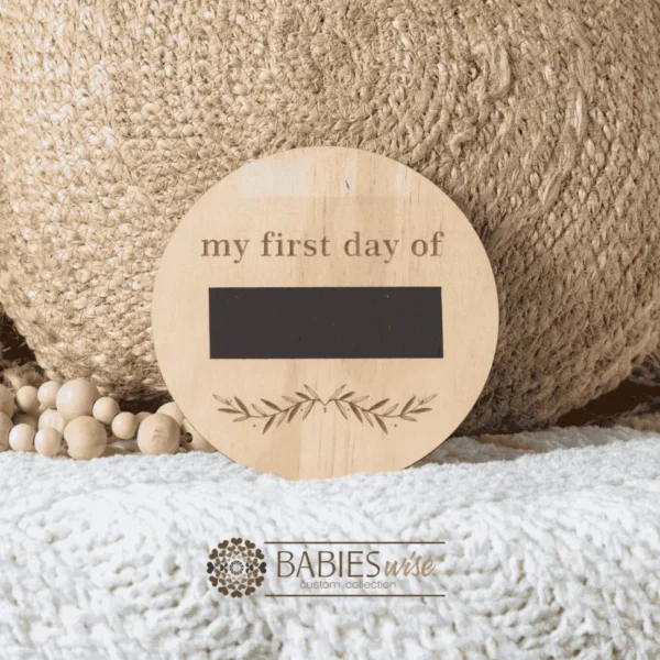Round timber "my first day of" milestone plaque with the words "my first day of Pre" engraved in dark letters. Styled on a soft white blanket with a woven pillow & wooden beads.Styled on a woven bassinett on a woven neutral mat.