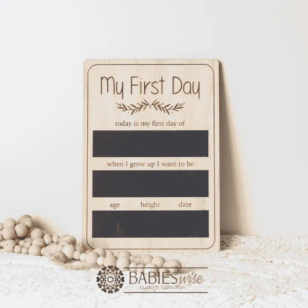 timber first day milestone board with reusable black writing panels and engraved prompts. Styled on a soft white blanket with wooden beads.