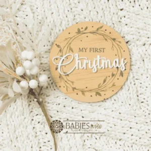 Round timber "My First Christmas" UV printed milestone plaque featuring a a delicate printed leaf wreath design with raised white acrylic "Christmas". Styled on a soft white blanket with a Christmas floral stem.