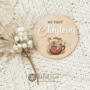 Round timber "My First Christmas" UV printed milestone plaque featuring a sweet koala wearing a red Santa hat sleeping in a santa sack with raised white acrylic "Christmas". Styled on a soft white blanket with a Christmas floral stem.