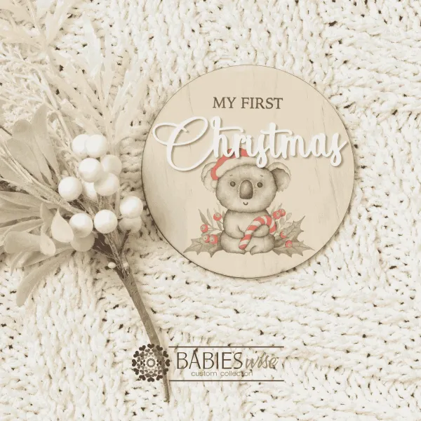 Round timber "My First Christmas" UV printed milestone plaque featuring a sweet koala wearing a red Santa hat holding a candy cane with raised white acrylic "Christmas". Styled on a soft white blanket with a Christmas floral stem.
