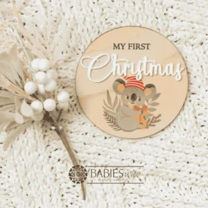 Round timber "My First Christmas" UV printed milestone plaque featuring a sweet koala wearing a red striped Santa hat holding a present with raised white acrylic "Christmas". Styled on a soft white blanket with a Christmas floral stem.