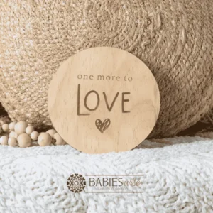 Round natural timber plaque engraved with words "One more to LOve" and a small heart illustration. Styled on a soft white blanket with a woven pillow & wooden beads.