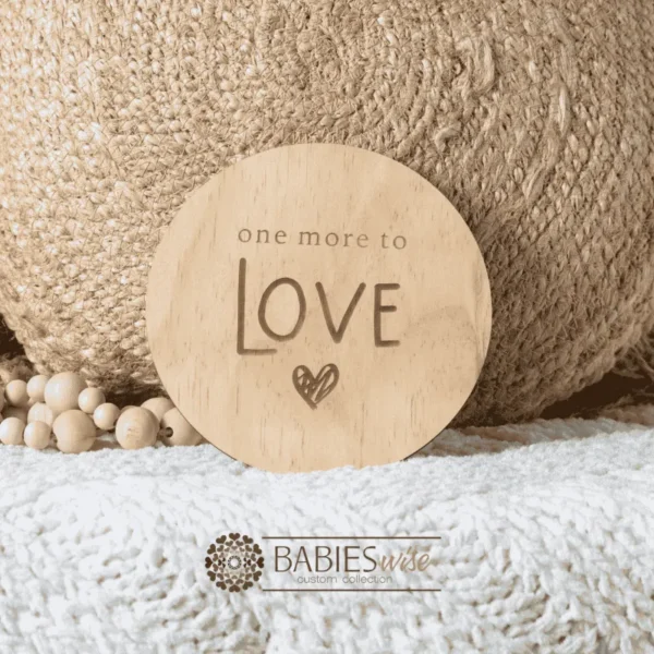 Round natural timber plaque engraved with words "One more to LOve" and a small heart illustration. Styled on a soft white blanket with a woven pillow & wooden beads.