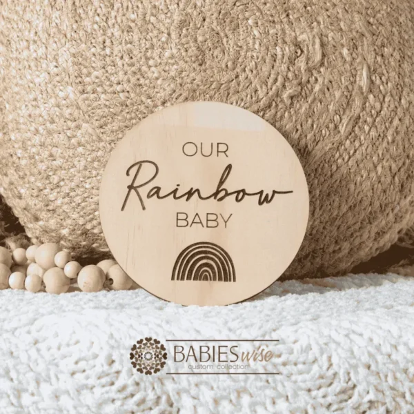 Round laser engraved timber plaque with the words "Our Rainbow Baby" in mixed script lettering and a small engraved rainbow illustration underneath. Styled on a soft white blanket with a woven pillow & wooden beads.
