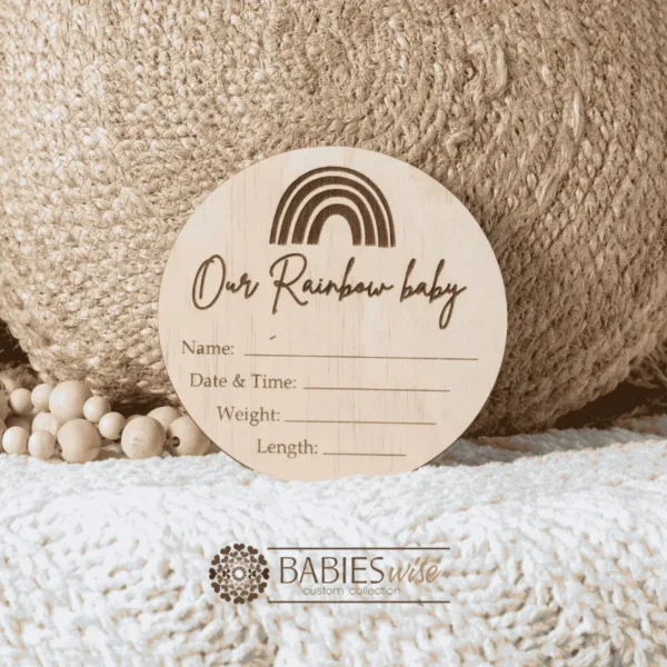 Round timber "Our Rainbow Baby" birth annoucement plaque with engraved rainbow and cursive lettering featuring spaces for baby's name, date and time, weight and lenght. Styled with a soft blanket and woven pillow.