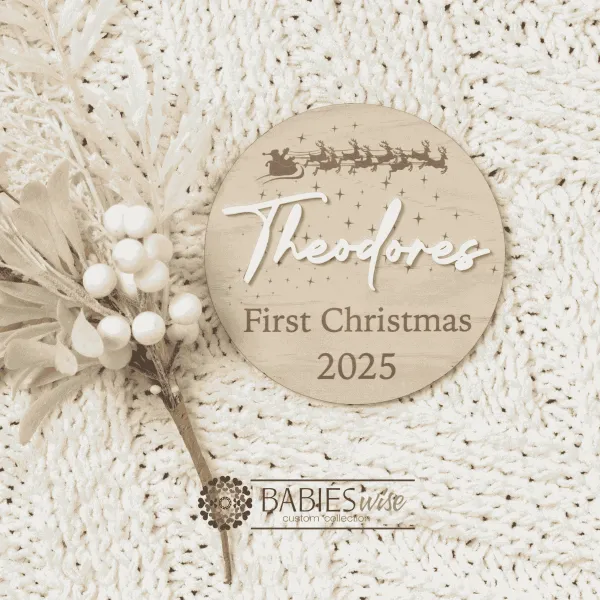 Round timber "My First Christmas" plaque featuring Santa's sleigh and reindeers flying across the night sky with stars and snowflakes with 3D acrylic child's name and printed year. Styled on a soft white blankt with a white Christmas wreath.