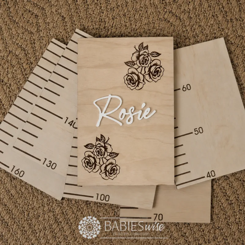 Personlised Timber Growth Charts laser engraved Header with roses and 3D acrylic child's name in script lettering. Styled as a flat-lay photo on a woven mat with the panels.