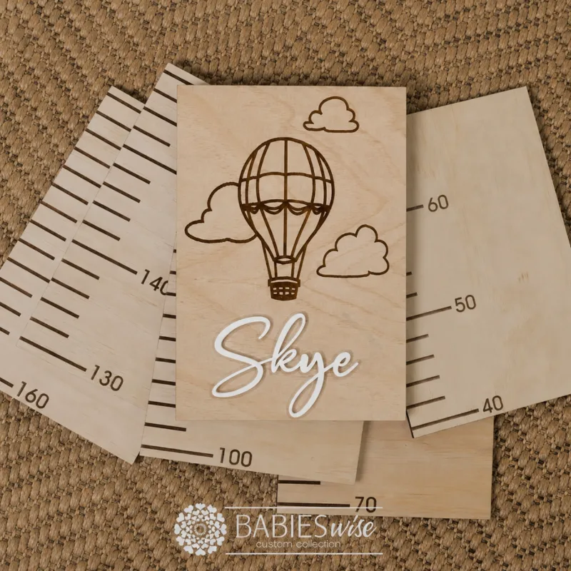 Personlised Timber Growth Charts laser engraved Header with a Hot Air Balloon and clouds with 3D acrylic child's name in script lettering. Styled as a flat-lay photo on a woven mat with the panels.