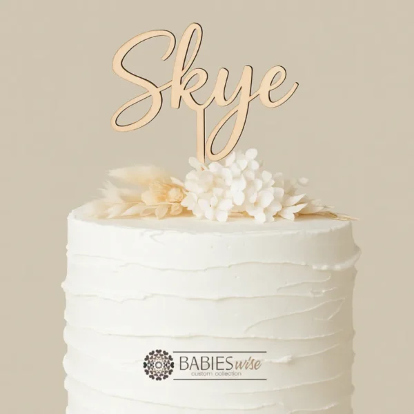 Personlised timber first cake topper featuring your child's name in a script lettering. Styled on a white cake with a floral arrangement.