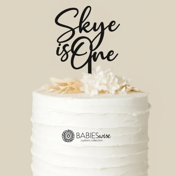 Personlised acrylic first birthday cake topper featuring your child's name in a script lettering above the words "is One" Styled on a white cake with a floral arrangement.