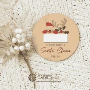 Round timber Christmas countdown plaque with a reusable white acrylic panel for writing the number of sleeps until Santa Claus visits. Available in two festive designs. Styled on a soft, white blanket.