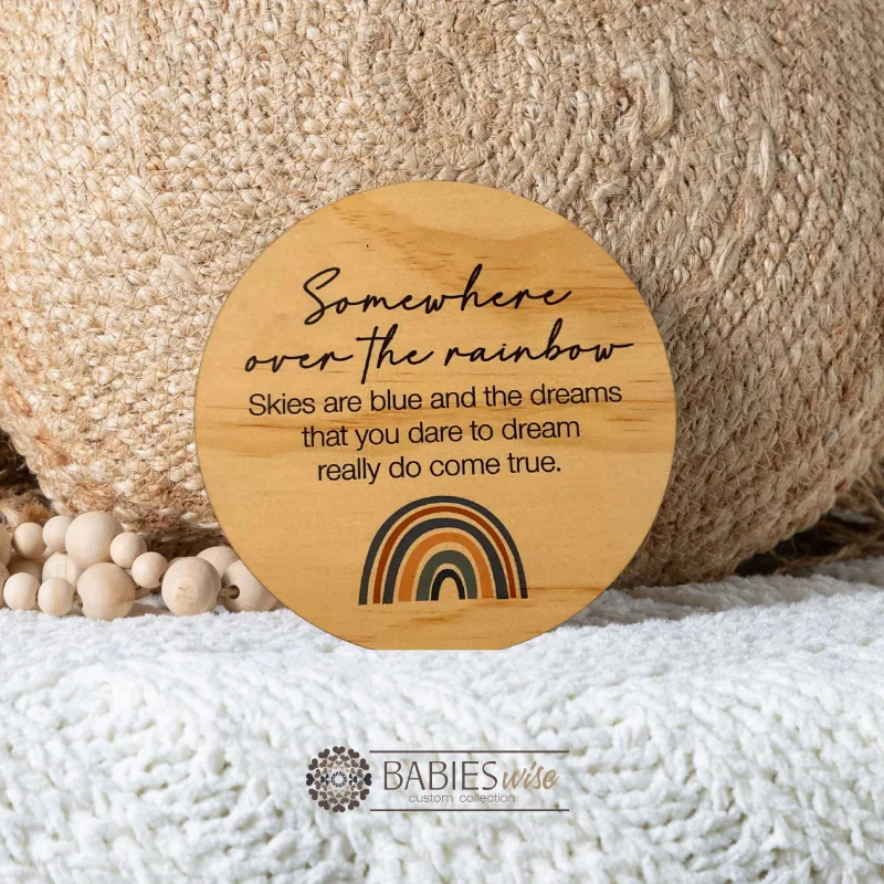 Round timber plaque with printed quote- "somewhere over the rainbow, Skies are blue and the dreams that you dare to dream really do come true". with a small brown-toned rainbow. Styled on a soft blanket withna woven pillow and wooden beads.