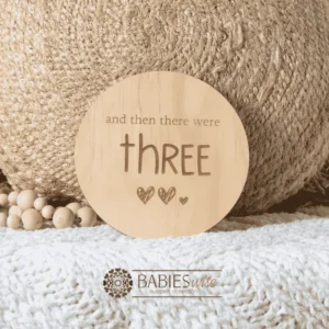 Round natural timber plaque engraved with words "and then there were three" and three small hearts illustration. Styled on a woven bassinett on a woven neutral mat.