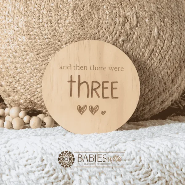 Round natural timber plaque engraved with words "and then there were three" and three small hearts illustration. Styled on a woven bassinett on a woven neutral mat.