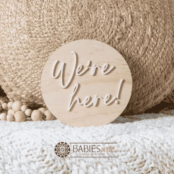 Round timber announcement plaque with raised blue acrylic script reading "We'ere Here!"Styled on a soft white blanket with a woven pillow & wooden beads.