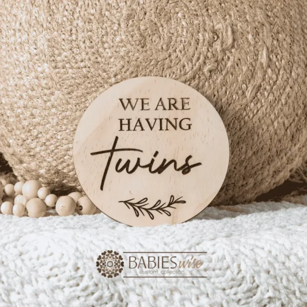 Round timber laser engraved plaque with wording "We are having Twins". Styled on a soft white blanket with a woven pillow & wooden beads.