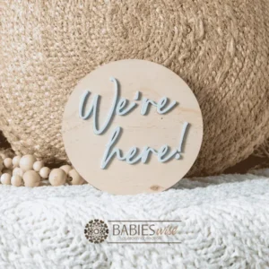 Round timber announcement plaque with raised blue acrylic script reading "We'ere Here!" Styled on a soft white blanket with a woven pillow & wooden beads.