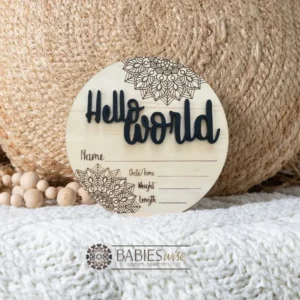Round timber Birth Announcement Plaque featuring Black raised acrylic "Hello World" lettering and engraved mandela floral designs, with spaces to record a baby's birth detals. Styled on a soft white blanket with a woven pillow & wooden beads.
