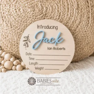 Round timber announcement plaque featuring the word "Introducing" above a personlised raised 3D acrylic name, with spaces below to record a baby's birth date, time, length, and weight. Styled on a soft white blanket with a woven pillow & wooden beads.