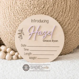 Round timber announcement plaque featuring the word "Introducing" above a personlised raised 3D acrylic name, with spaces below to record a baby's birth date, time, length, and weight. Styled on a soft white blanket with a woven pillow & wooden beads.