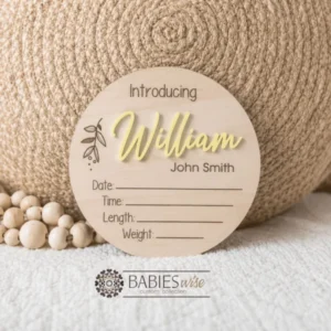 Round timber announcement plaque featuring the word "Introducing" above a personlised raised 3D acrylic name, with spaces below to record a baby's birth date, time, length, and weight. Styled on a soft white blanket with a woven pillow & wooden beads.