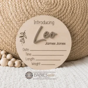 Round timber announcement plaque featuring the word "Introducing" above a personlised raised 3D acrylic name, with spaces below to record a baby's birth date, time, length, and weight. Styled on a soft white blanket with a woven pillow & wooden beads.