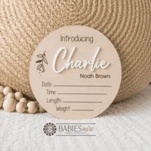 Round timber announcement plaque featuring the word "Introducing" above a personlised raised 3D acrylic name, with spaces below to record a baby's birth date, time, length, and weight. Styled on a soft white blanket with a woven pillow & wooden beads.