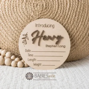 Round timber announcement plaque featuring the word "Introducing" above a personlised raised 3D acrylic name, with spaces below to record a baby's birth date, time, length, and weight. Styled on a soft white blanket with a woven pillow & wooden beads.