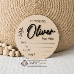 Round timber announcement plaque featuring the word "Introducing" above a personlised raised 3D acrylic name, with spaces below to record a baby's birth date, time, length, and weight. Styled on a soft white blanket with a woven pillow & wooden beads.