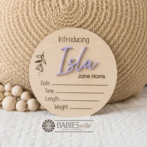 Round timber announcement plaque featuring the word "Introducing" above a personlised raised 3D acrylic name, with spaces below to record a baby's birth date, time, length, and weight. Styled on a soft white blanket with a woven pillow & wooden beads.
