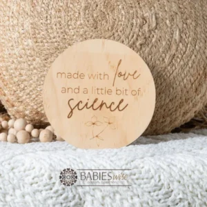 Round timber plaque engraved with the words "made with love and a little bit of science" in mixed fonts with a small floral line drawing at the bottom. Styled with a soft blanket and set in a frame with the baby's feet.