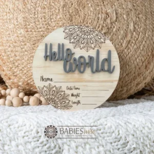 Round timber Birth Announcement Plaque featuring coffee raised acrylic "Hello World" lettering and engraved mandela floral designs, with spaces to record a baby's birth detals. Styled on a soft white blanket with a woven pillow & wooden beads.