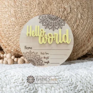 Round timber Birth Announcement Plaque featuring yellow raised acrylic "Hello World" lettering and engraved mandela floral designs, with spaces to record a baby's birth detals. Styled on a soft white blanket with a woven pillow & wooden beads.