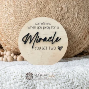 Round timber twin pregnancy announcement plaque featuring the phrase :sometimes when you pray for a Miracle, you get two" with the word "Miracle" in raised 3D acrylic . Personlised with the surname, month and year UV printed along the bottom of the plaque. Styled on a soft white blanket with a woven pillow & wooden beads.