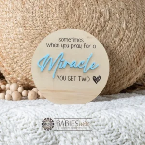 Round timber twin pregnancy announcement plaque featuring the phrase :sometimes when you pray for a Miracle, you get two" with the word "Miracle" in raised 3D acrylic . Personlised with the surname, month and year UV printed along the bottom of the plaque. Styled on a soft white blanket with a woven pillow & wooden beads.